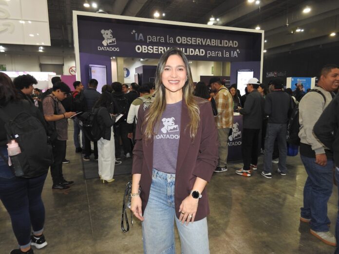 Fotografía en plano medio de Queennie López, Senior Alliances Manager para Datadog en México, sonriendo frente al estand de la marca en un evento tecnológico con el lema 