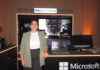 Dell Technologies y la revolución empresarial: inteligencia artificial a la medida con AMD Fotografía de plano medio de Margarita Gutiérrez posando sonriente en traje ejecutivo verde claro frente a un stand de exhibición tecnológica iluminado. Sobre la barra del stand se observa una computadora portátil y un monitor empresarial encendidos, acompañados por el letrero de Dell Technologies en la parte superior y el logotipo de Microsoft en el panel frontal inferior.