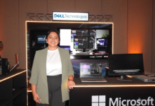 Dell Technologies y la revolución empresarial: inteligencia artificial a la medida con AMD Fotografía de plano medio de Margarita Gutiérrez posando sonriente en traje ejecutivo verde claro frente a un stand de exhibición tecnológica iluminado. Sobre la barra del stand se observa una computadora portátil y un monitor empresarial encendidos, acompañados por el letrero de Dell Technologies en la parte superior y el logotipo de Microsoft en el panel frontal inferior.