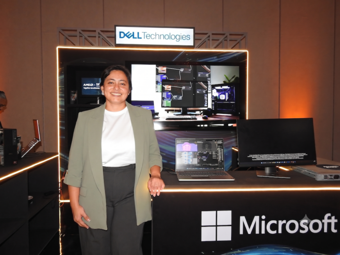 Sinergia estratégica entre Dell Technologies y AMD para el cómputo de inteligencia artificial Fotografía de plano medio de Margarita Gutiérrez posando sonriente en traje ejecutivo verde claro frente a un stand de exhibición tecnológica iluminado. Sobre la barra del stand se observa una computadora portátil y un monitor empresarial encendidos, acompañados por el letrero de Dell Technologies en la parte superior y el logotipo de Microsoft en el panel frontal inferior.