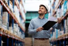 Radii: Manufactura mexicana pierde proyectos globales por falta de presencia digital Fotografía de un hombre sonriendo dentro de un almacén con pasillos de estantería industrial de fondo. El sujeto viste una camisa azul, pantalones color caqui y un casco de seguridad blanco, mientras sostiene una tabla con pinza y revisa un documento impreso.