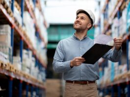 Radii: Manufactura mexicana pierde proyectos globales por falta de presencia digital Fotografía de un hombre sonriendo dentro de un almacén con pasillos de estantería industrial de fondo. El sujeto viste una camisa azul, pantalones color caqui y un casco de seguridad blanco, mientras sostiene una tabla con pinza y revisa un documento impreso.