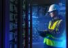 ABB: La orquestación energética marcará el futuro de la competitividad industrial Fotografía en plano medio de un ingeniero de campo con casco de seguridad blanco, chaleco de alta visibilidad y lentes de protección, revisando diagnósticos en una computadora portátil dentro de un centro de datos iluminado con luces azules.