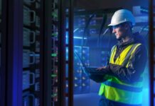 ABB: La orquestación energética marcará el futuro de la competitividad industrial Fotografía en plano medio de un ingeniero de campo con casco de seguridad blanco, chaleco de alta visibilidad y lentes de protección, revisando diagnósticos en una computadora portátil dentro de un centro de datos iluminado con luces azules.