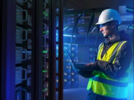 ABB: La orquestación energética marcará el futuro de la competitividad industrial Fotografía en plano medio de un ingeniero de campo con casco de seguridad blanco, chaleco de alta visibilidad y lentes de protección, revisando diagnósticos en una computadora portátil dentro de un centro de datos iluminado con luces azules.