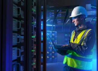 ABB: La orquestación energética marcará el futuro de la competitividad industrial Fotografía en plano medio de un ingeniero de campo con casco de seguridad blanco, chaleco de alta visibilidad y lentes de protección, revisando diagnósticos en una computadora portátil dentro de un centro de datos iluminado con luces azules.