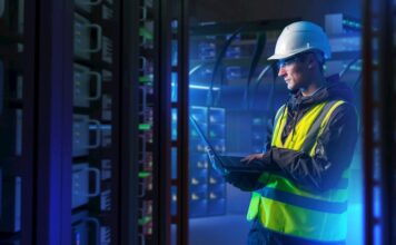 ABB: La orquestación energética marcará el futuro de la competitividad industrial Fotografía en plano medio de un ingeniero de campo con casco de seguridad blanco, chaleco de alta visibilidad y lentes de protección, revisando diagnósticos en una computadora portátil dentro de un centro de datos iluminado con luces azules.