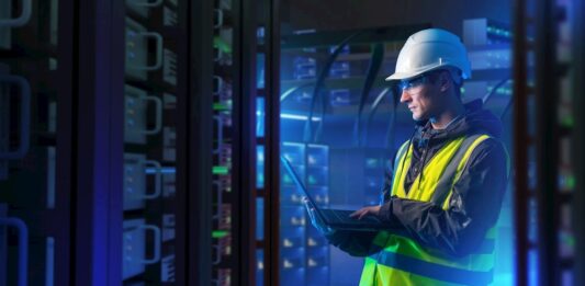 ABB: La orquestación energética marcará el futuro de la competitividad industrial Fotografía en plano medio de un ingeniero de campo con casco de seguridad blanco, chaleco de alta visibilidad y lentes de protección, revisando diagnósticos en una computadora portátil dentro de un centro de datos iluminado con luces azules.