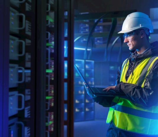 ABB: La orquestación energética marcará el futuro de la competitividad industrial Fotografía en plano medio de un ingeniero de campo con casco de seguridad blanco, chaleco de alta visibilidad y lentes de protección, revisando diagnósticos en una computadora portátil dentro de un centro de datos iluminado con luces azules.