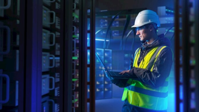 Un especialista en electrificación supervisa la continuidad operativa de la infraestructura eléctrica en un entorno de servidores. Fotografía en plano medio de un ingeniero de campo con casco de seguridad blanco, chaleco de alta visibilidad y lentes de protección, revisando diagnósticos en una computadora portátil dentro de un centro de datos iluminado con luces azules.