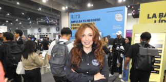 El futuro del sector enterprise está en la nube y la inteligencia artificial junto a Itera Fotografía en plano medio de Alondra Castro, Key Account Manager en Itera, sonriendo con los brazos cruzados en una camisa negra corporativa y jeans azules, frente al estand de la empresa en una convención tecnológica con el mensaje "IMPULSA TU NEGOCIO CON IA" al fondo.