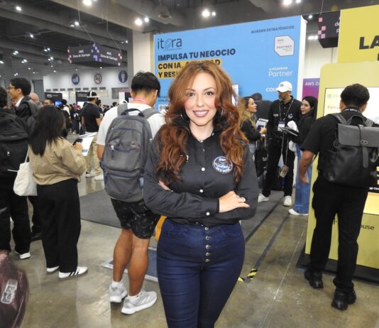 El futuro del sector enterprise está en la nube y la inteligencia artificial junto a Itera Fotografía en plano medio de Alondra Castro, Key Account Manager en Itera, sonriendo con los brazos cruzados en una camisa negra corporativa y jeans azules, frente al estand de la empresa en una convención tecnológica con el mensaje "IMPULSA TU NEGOCIO CON IA" al fondo.