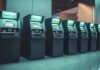 Banca omnicanal con Auriga: la ventaja competitiva de conectar todos los canales Fotografía en plano general de una hilera de cajeros automáticos multifuncionales iluminados con luz verde en el interior de una sucursal bancaria moderna y pulcra.