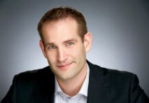 Fortinet: Construir una cultura que priorice la seguridad ante un panorama de amenazas en aceleración Fotografía de retrato en plano medio de Derek Manky, Vicepresidente Global de Inteligencia de Amenazas en Fortinet, vistiendo saco oscuro sobre camisa a cuadros, sonriendo frente a un fondo neutro de estudio.