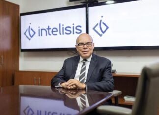 Intelisis: La evolución del ERP; de la gestión de información a la toma de decisiones Fotografía de retrato corporativo en plano medio de Dionisio Castillo Guillén, Director General de Intelisis Software, vistiendo traje oscuro con corbata a rayas y lentes, sentado a la mesa de una sala de juntas frente a dos pantallas con el logotipo de Intelisis.