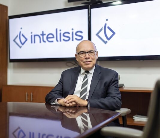 Intelisis: La evolución del ERP; de la gestión de información a la toma de decisiones Fotografía de retrato corporativo en plano medio de Dionisio Castillo Guillén, Director General de Intelisis Software, vistiendo traje oscuro con corbata a rayas y lentes, sentado a la mesa de una sala de juntas frente a dos pantallas con el logotipo de Intelisis.