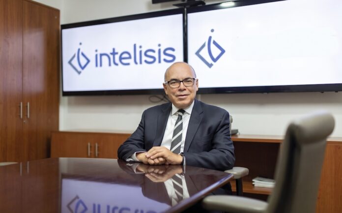 Fotografía de retrato corporativo en plano medio de Dionisio Castillo Guillén, Director General de Intelisis Software, vistiendo traje oscuro con corbata a rayas y lentes, sentado a la mesa de una sala de juntas frente a dos pantallas con el logotipo de Intelisis.