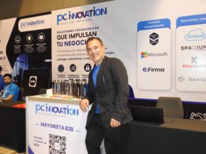 Fotografía en plano medio de Eleazar Carrillo, Director General en corporativo PC Software, vistiendo saco oscuro sobre playera azul, apoyado en el estand promocional de "PC Innovation - Mayorista B2B".