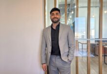 Streambe: Equipos más chicos y proyectos más baratos: el nuevo paradigma de programación que impulsa la IA Fotografía de retrato corporativo en plano medio de Gastón Gugliotta, CEO de Streambe, vistiendo traje gris claro sobre camisa negra abierta, posando sonriente con una mano en el bolsillo frente a un cancel divisorio de madera y cristal en una oficina moderna.