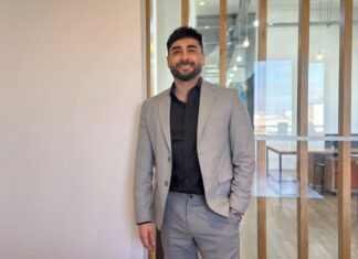 Streambe: Equipos más chicos y proyectos más baratos: el nuevo paradigma de programación que impulsa la IA Fotografía de retrato corporativo en plano medio de Gastón Gugliotta, CEO de Streambe, vistiendo traje gris claro sobre camisa negra abierta, posando sonriente con una mano en el bolsillo frente a un cancel divisorio de madera y cristal en una oficina moderna.