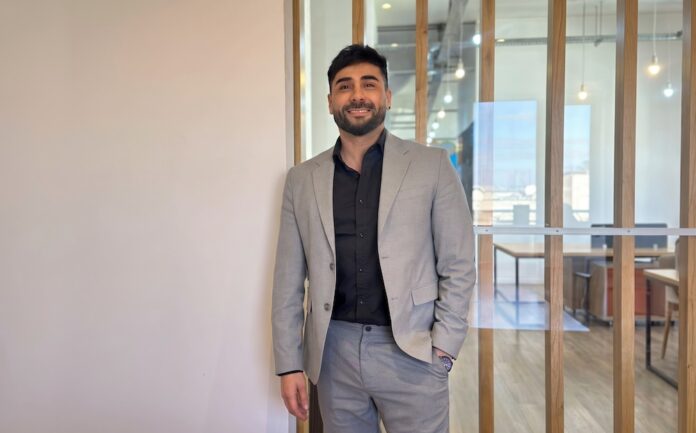 Fotografía de retrato corporativo en plano medio de Gastón Gugliotta, CEO de Streambe, vistiendo traje gris claro sobre camisa negra abierta, posando sonriente con una mano en el bolsillo frente a un cancel divisorio de madera y cristal en una oficina moderna.