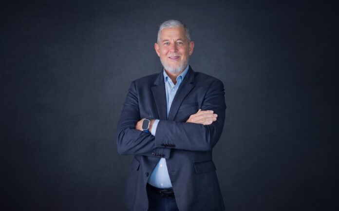 Fotografía de retrato corporativo en plano medio de Max Mayorga, Vicepresidente para América Latina de Ivanti, vistiendo saco azul marino sobre camisa azul claro, posando sonriente con los brazos cruzados frente a un fondo gris neutro.