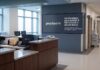 Proofpoint alerta que los ciberataques interrumpen la atención médica en el 74% de las clínicas de EE. UU. Fotografía en plano general del pasillo y recepción de un hospital con mostradores de enfermería desiertos, monitores de computadora que muestran mensajes de sistema fuera de línea y una pantalla institucional en el muro con el logotipo de Proofpoint y la leyenda "Los ciberataques ponen en riesgo la continuidad de la atención médica".