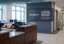 Proofpoint alerta que los ciberataques interrumpen la atención médica en el 74% de las clínicas de EE. UU. Fotografía en plano general del pasillo y recepción de un hospital con mostradores de enfermería desiertos, monitores de computadora que muestran mensajes de sistema fuera de línea y una pantalla institucional en el muro con el logotipo de Proofpoint y la leyenda "Los ciberataques ponen en riesgo la continuidad de la atención médica".