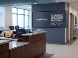 Proofpoint alerta que los ciberataques interrumpen la atención médica en el 74% de las clínicas de EE. UU. Fotografía en plano general del pasillo y recepción de un hospital con mostradores de enfermería desiertos, monitores de computadora que muestran mensajes de sistema fuera de línea y una pantalla institucional en el muro con el logotipo de Proofpoint y la leyenda "Los ciberataques ponen en riesgo la continuidad de la atención médica".