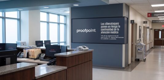Proofpoint alerta que los ciberataques interrumpen la atención médica en el 74% de las clínicas de EE. UU. Fotografía en plano general del pasillo y recepción de un hospital con mostradores de enfermería desiertos, monitores de computadora que muestran mensajes de sistema fuera de línea y una pantalla institucional en el muro con el logotipo de Proofpoint y la leyenda "Los ciberataques ponen en riesgo la continuidad de la atención médica".