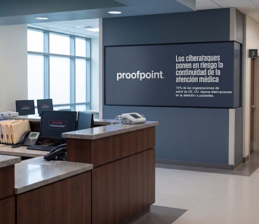 Proofpoint alerta que los ciberataques interrumpen la atención médica en el 74% de las clínicas de EE. UU. Fotografía en plano general del pasillo y recepción de un hospital con mostradores de enfermería desiertos, monitores de computadora que muestran mensajes de sistema fuera de línea y una pantalla institucional en el muro con el logotipo de Proofpoint y la leyenda "Los ciberataques ponen en riesgo la continuidad de la atención médica".