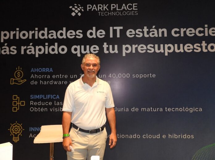 Fotografía en plano medio de Rogelio Moncada, Director Comercial para North Latam en Park Place Technologies, vistiendo camisa blanca tipo polo corporativa, posando sonriente frente a un muro publicitario con la frase 