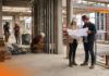 Cómo GAYA impulsa la IA y la gestión digital para anticipar sobrecostos en la construcción Fotografía en plano entero de dos ingenieros en obra revisando planos en papel con cascos de seguridad, mientras otros trabajadores realizan labores de construcción al fondo dentro de una estructura de concreto.