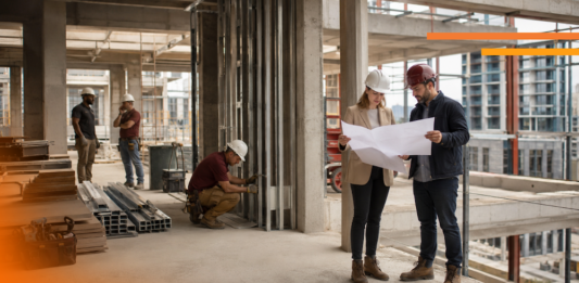 Cómo GAYA impulsa la IA y la gestión digital para anticipar sobrecostos en la construcción Fotografía en plano entero de dos ingenieros en obra revisando planos en papel con cascos de seguridad, mientras otros trabajadores realizan labores de construcción al fondo dentro de una estructura de concreto.