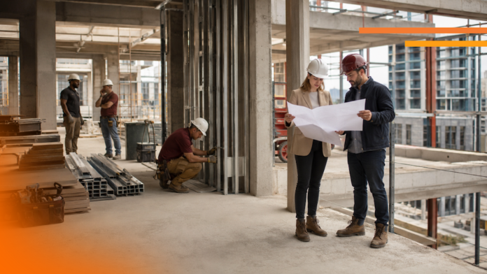 Supervisores e ingenieros de obra revisan planes de construcción para mitigar riesgos de sobrecosto Fotografía en plano entero de dos ingenieros en obra revisando planos en papel con cascos de seguridad, mientras otros trabajadores realizan labores de construcción al fondo dentro de una estructura de concreto.